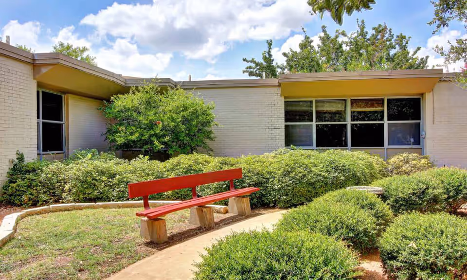 Small courtyard outside a low brick building with a red bench, trimmed bushes, and large windows under a partly cloudy sky.