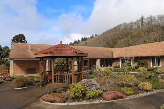 Outdoor courtyard area of a senior living facility with a wooden gazebo surrounded by landscaped garden beds and shrubs, single-story building with brown roof in the background, and wooded hillside under a partly cloudy sky.