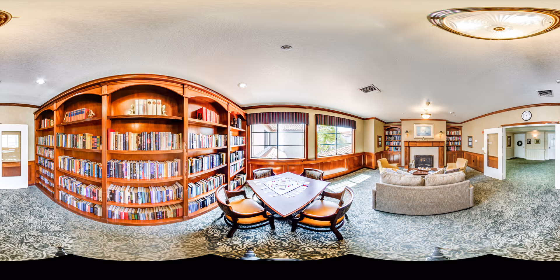 A spacious and well-lit library or common room with large wooden bookshelves filled with books along one wall. In the center, there is a square table with four chairs around it, and a game or puzzle is set up on the table. The room features a comfortable seating area with a sofa and armchairs arranged around a fireplace. Large windows with valances allow natural light to fill the room, and the floor is covered with a patterned carpet. The walls have wood paneling and there is a clock above a doorway leading to another room.