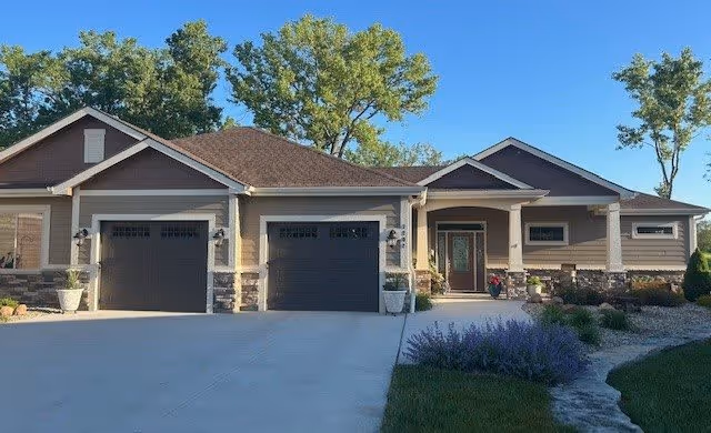 Front exterior view of a single-story residential building with a three-car garage, stone and siding facade, a covered entrance with columns, and landscaping including bushes and trees under a clear blue sky.