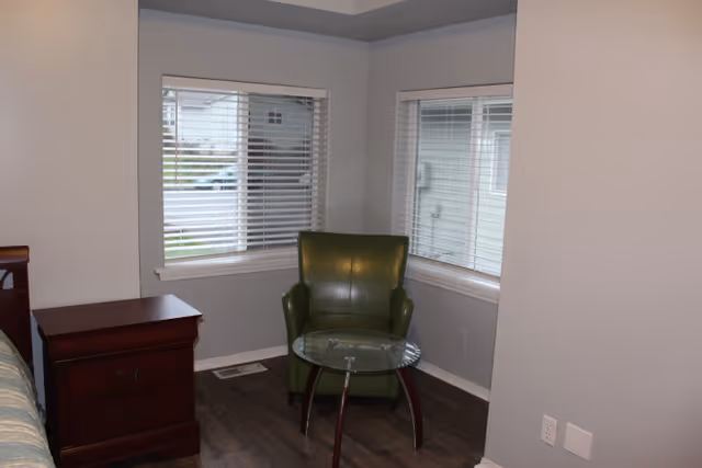 A corner of a bedroom with a green armchair, a glass side table, a wooden nightstand, and two windows with blinds.