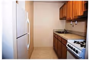 A small kitchen with wooden cabinets, a white refrigerator on the left, a countertop with a sink, and a gas stove on the right side.