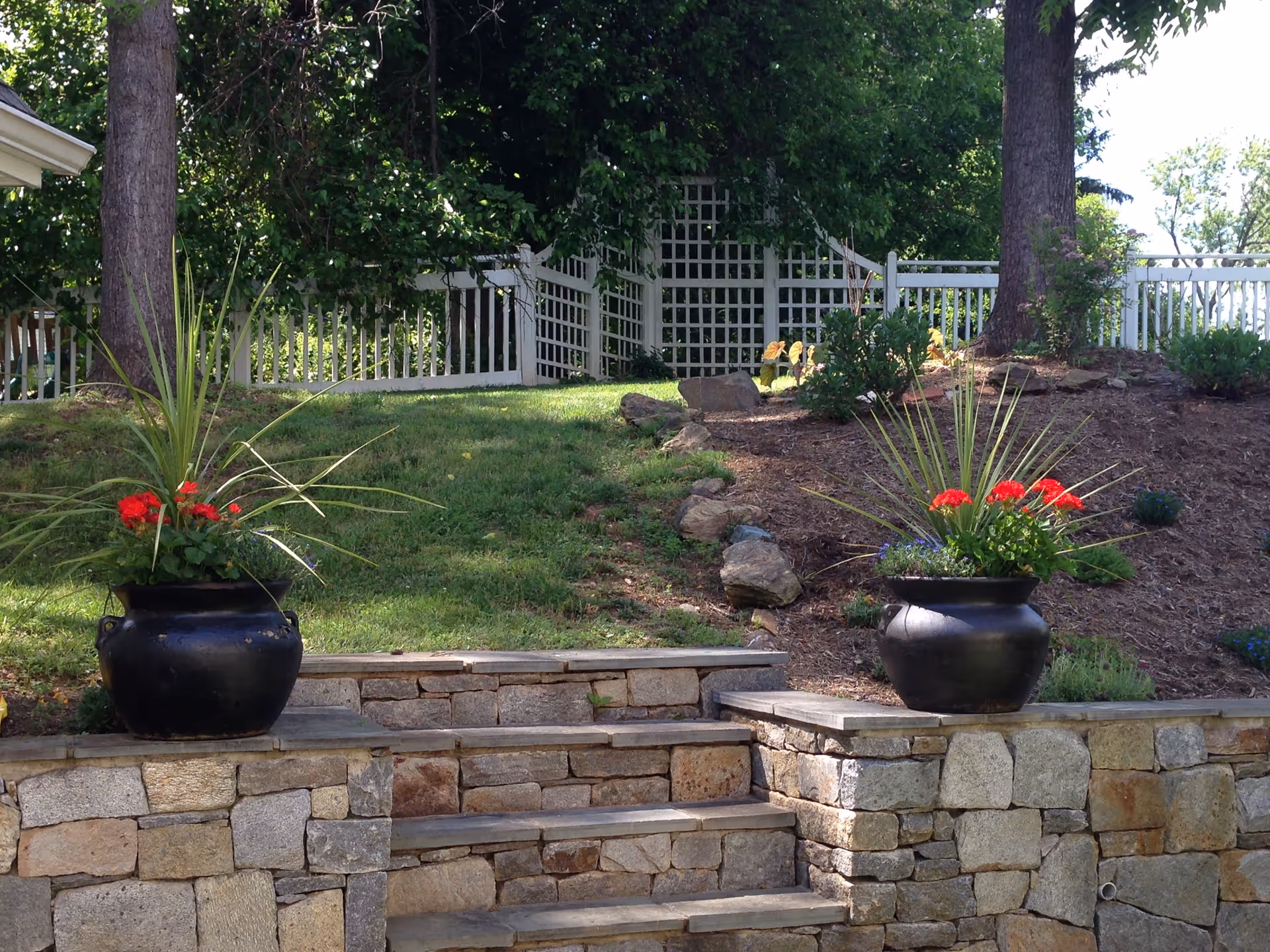 Stone steps leading up to a grassy area with two large black planters on either side, each containing green plants and red flowers. In the background, there is a white wooden fence and trellis with trees and shrubs surrounding the area.