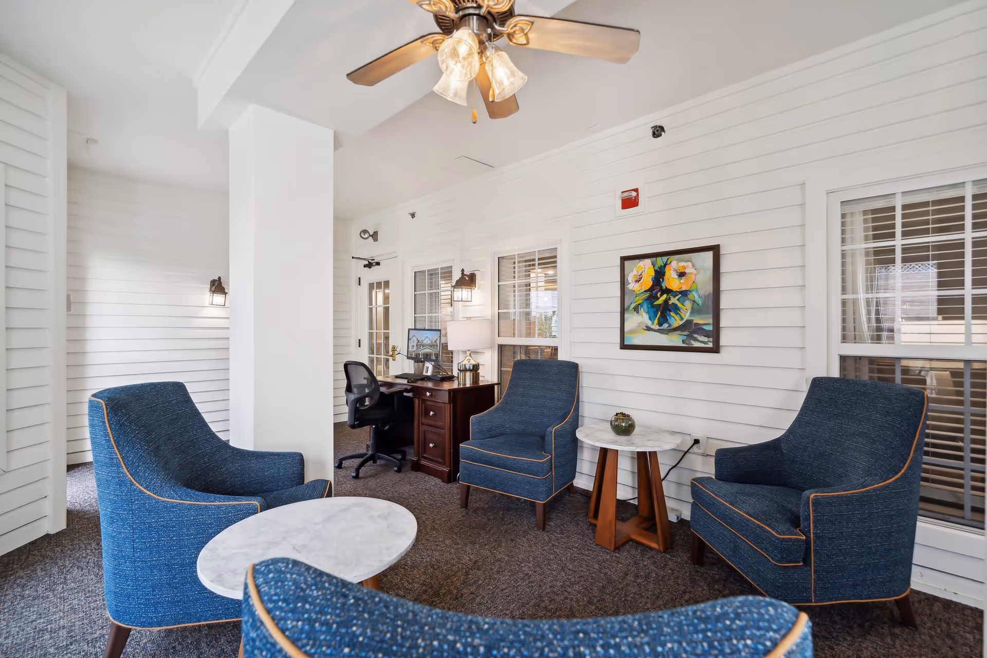 A bright indoor lounge with blue upholstered armchairs, marble-top side tables, a desk, ceiling fan, and framed wall art.