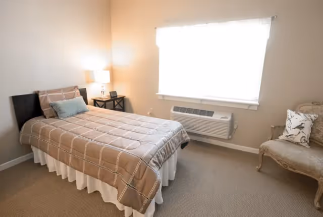 Simple furnished bedroom with a single bed, bedside table and lamp, a bright window with an air-conditioning unit, and an upholstered chair.