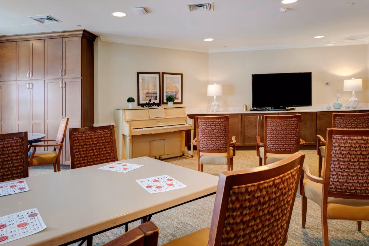 Communal activity room with tables and chairs facing a TV, a piano against the wall, and built-in cabinets.