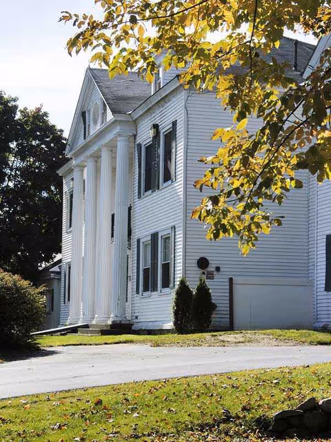 White two-story building with tall columns and a front entrance, set on a green lawn.