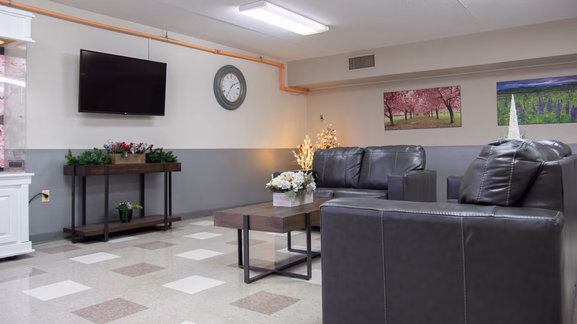 A lounge area with black leather sofas and a wooden coffee table with a flower arrangement. A flat-screen TV is mounted on the wall above a console table with plants. The walls are painted gray and white, decorated with landscape paintings and a round clock. The floor has a checkered tile pattern.