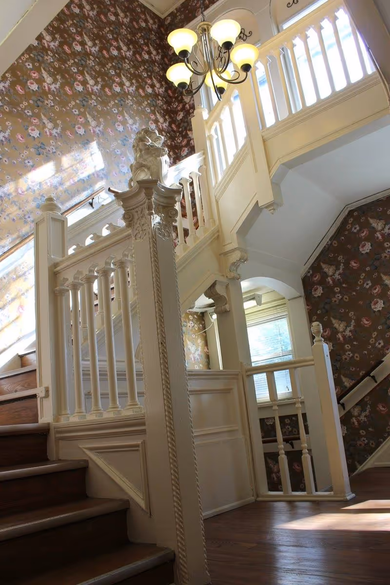 Interior view of a staircase with white wooden railings and newel posts, featuring a decorative lion sculpture on top of one post. The walls are covered with floral wallpaper, and a chandelier with five lights hangs from the ceiling. Sunlight streams through a window, casting shadows on the wall and floor.