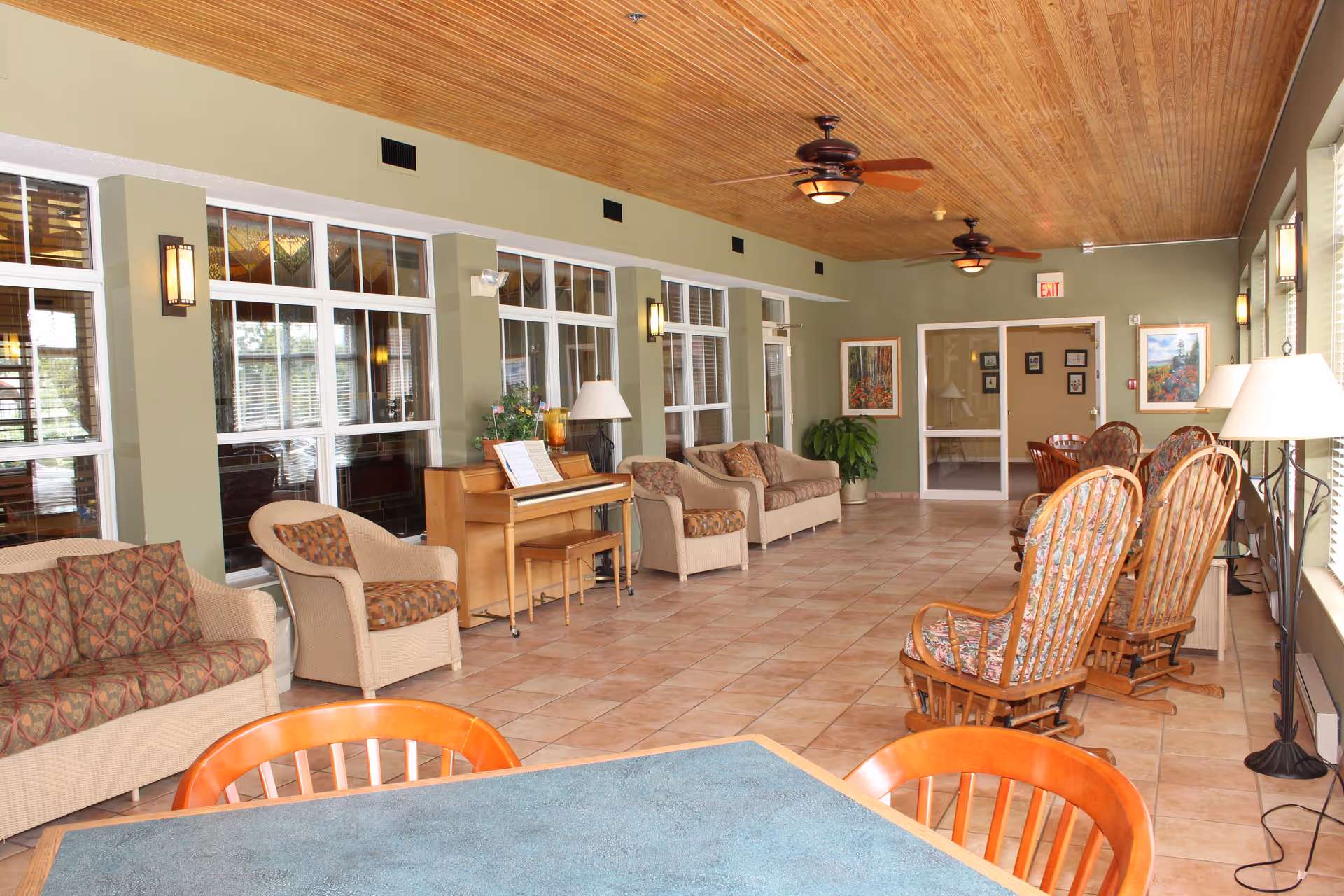 A spacious common area with tiled floor and wooden ceiling featuring ceiling fans. The room has multiple seating options including cushioned wicker sofas and chairs, wooden rocking chairs with floral cushions, and a table with chairs in the foreground. There is a piano against one wall with sheet music on it, lamps, potted plants, and framed artwork on the walls. Large windows line one side of the room, letting in natural light.