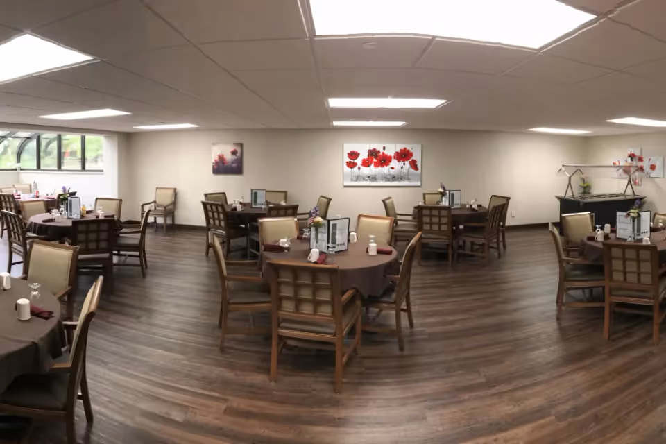 A spacious dining room with several round tables covered with brown tablecloths, each surrounded by wooden chairs with cushioned seats. The tables are set with menus, napkins, and small decorative items. The room has wood flooring, white walls, and ceiling lights. There are paintings of red flowers on the walls and a window on the left side letting in natural light.