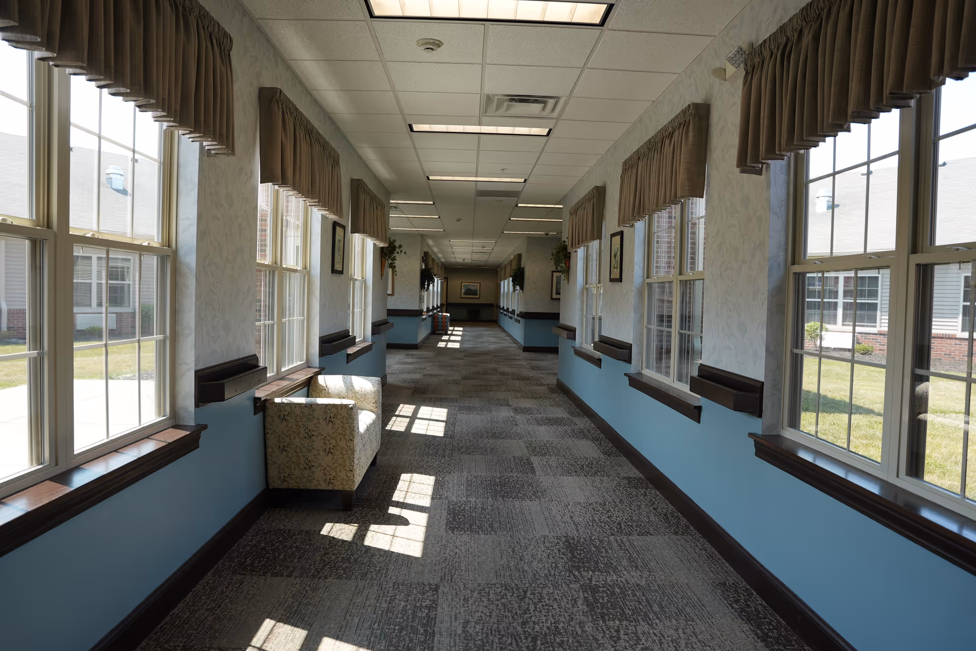 A long, well-lit hallway with large windows on both sides, covered with beige valance curtains. The walls are painted light blue on the lower half and patterned wallpaper on the upper half. There is a single floral armchair placed near the windows on the left side. The carpet has a patterned design, and the ceiling has recessed lighting panels. Outside the windows, a grassy courtyard and parts of the building exterior are visible.