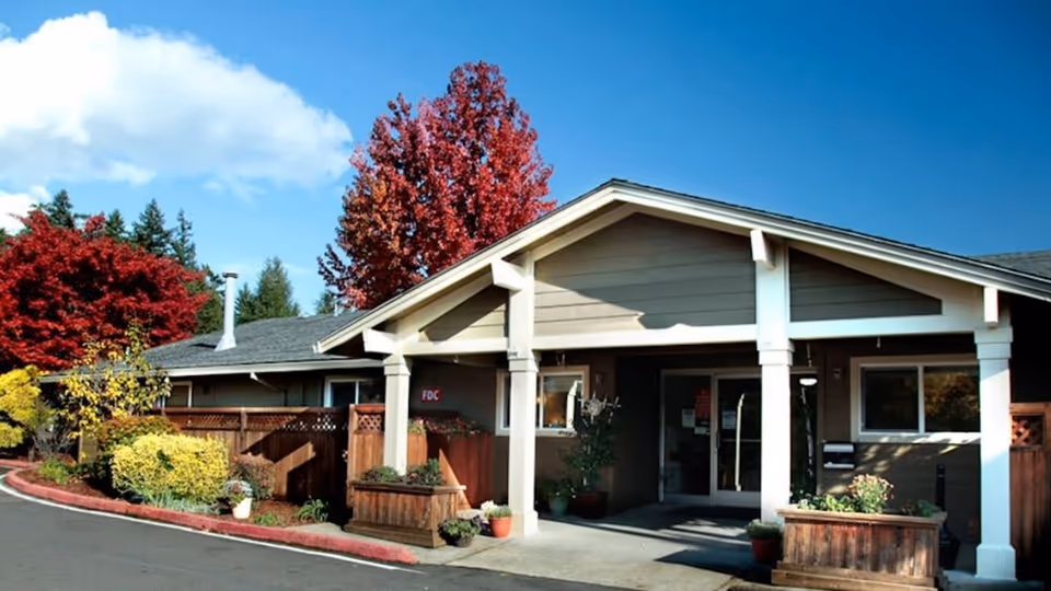 Front entrance of a single-story care facility with a covered porch, planters, and colorful autumn trees.