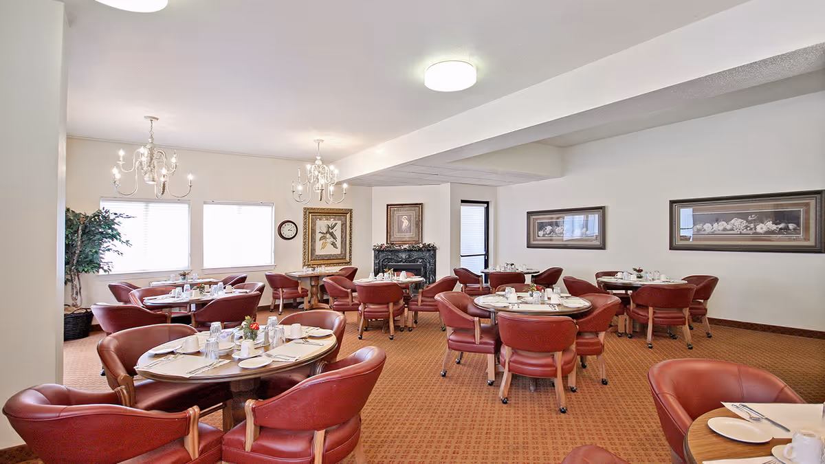 Spacious dining room with round tables set for meals and red upholstered chairs under chandeliers.
