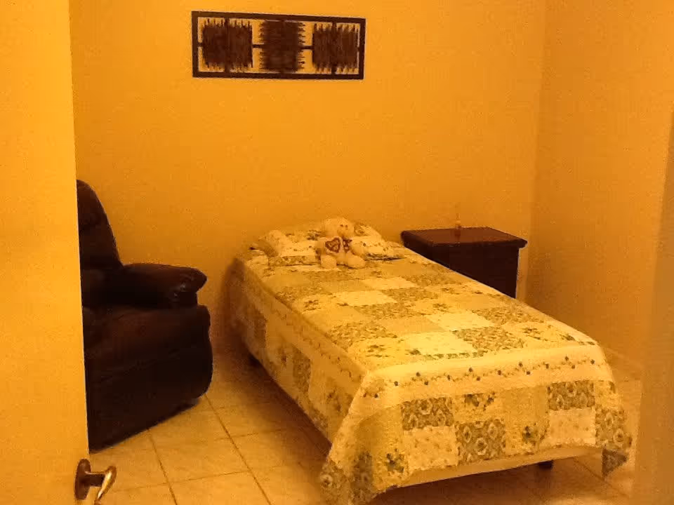 A small bedroom with a single bed covered in a patchwork quilt, a teddy bear placed on the bed, a dark armchair to the left, a wooden nightstand to the right, and a decorative wall hanging above the bed.