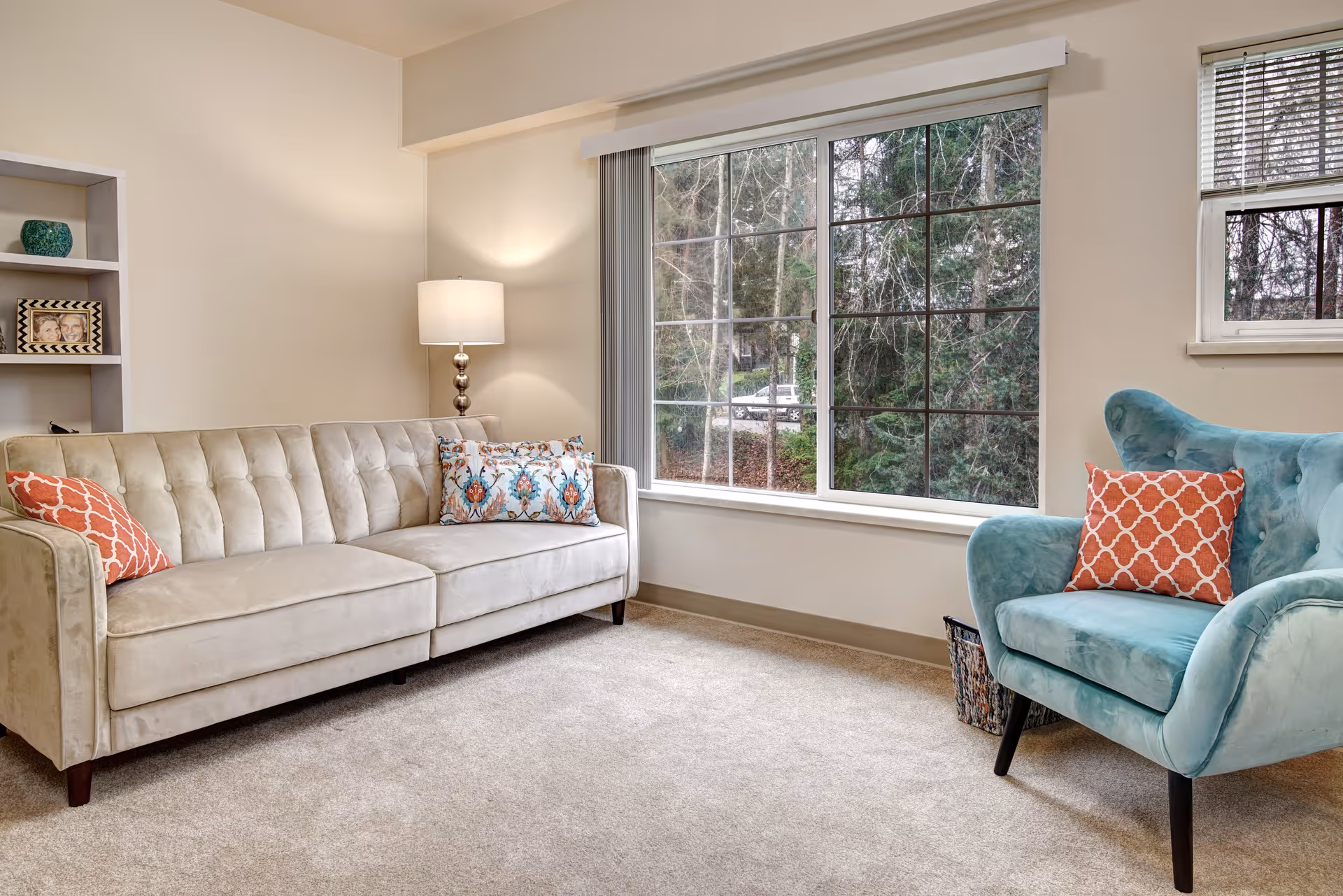 A cozy living room with a beige tufted sofa adorned with patterned pillows, a teal armchair with an orange patterned pillow, a floor lamp with a white shade, a large window showing trees outside, and a small built-in shelf with a framed photo and decorative items.