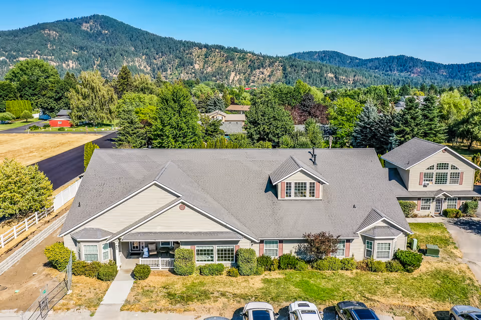 Aerial view of a single-story senior living facility building with a gray roof and beige siding, surrounded by green bushes and trees. There are several cars parked in front of the building, and a mountainous landscape is visible in the background under a clear blue sky.