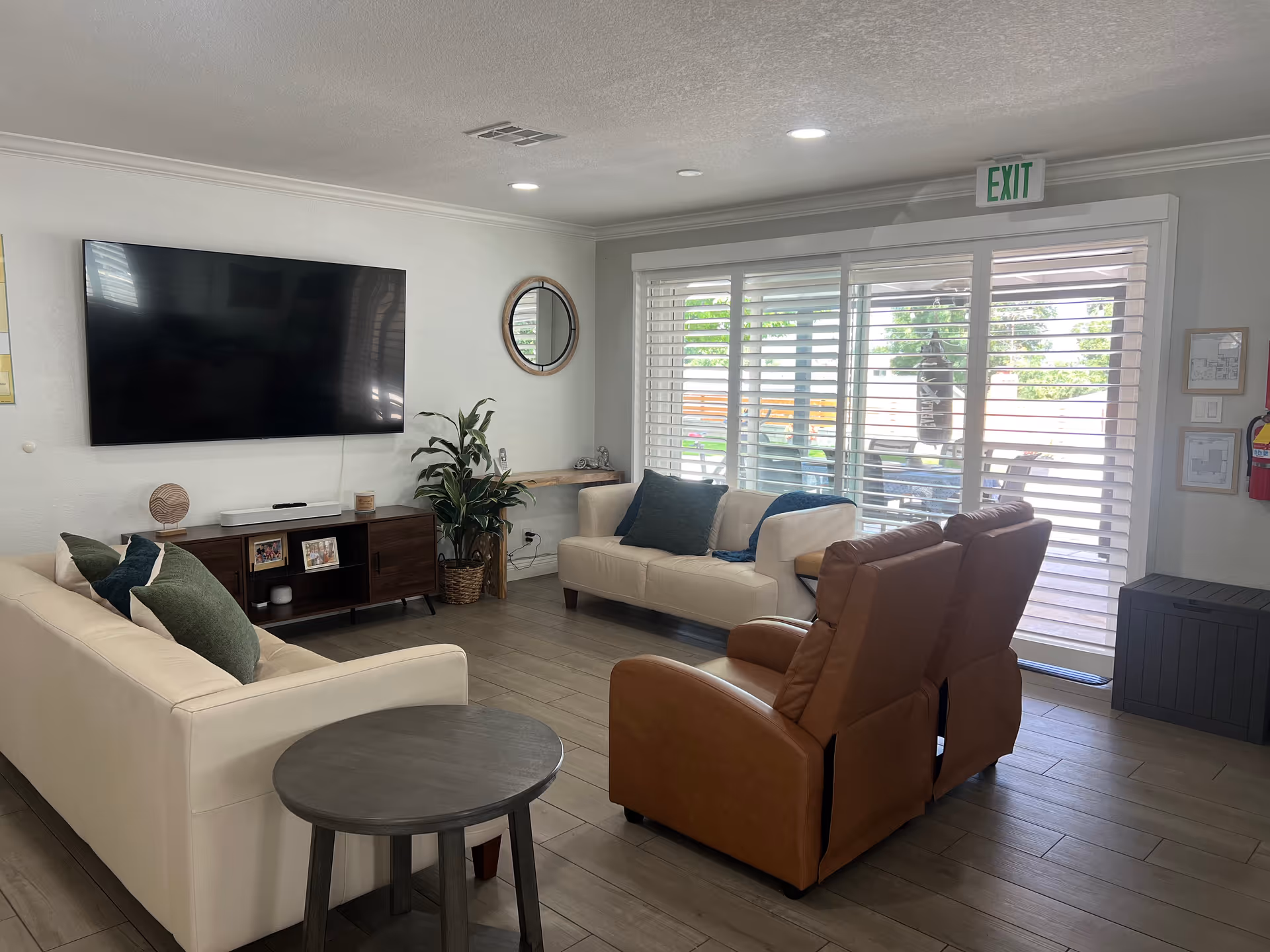 A cozy living room with two cream-colored sofas, two brown recliner chairs, a round side table, a large flat-screen TV mounted on the wall, a wooden TV stand with framed photos and decor, a potted plant, a round wall mirror, and large windows with white blinds letting in natural light. An exit sign is visible above the sliding glass door leading outside.