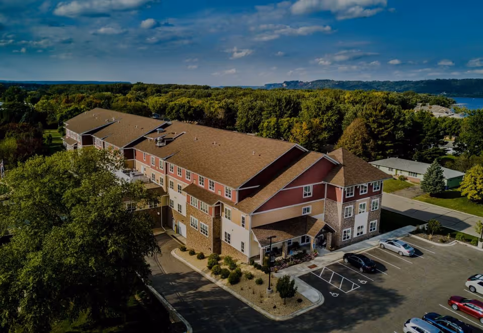 Aerial view of a large senior living facility building with a brown roof and red and beige exterior walls, surrounded by trees and greenery. The building has a parking lot with several cars parked, and a clear blue sky with some clouds is visible above.