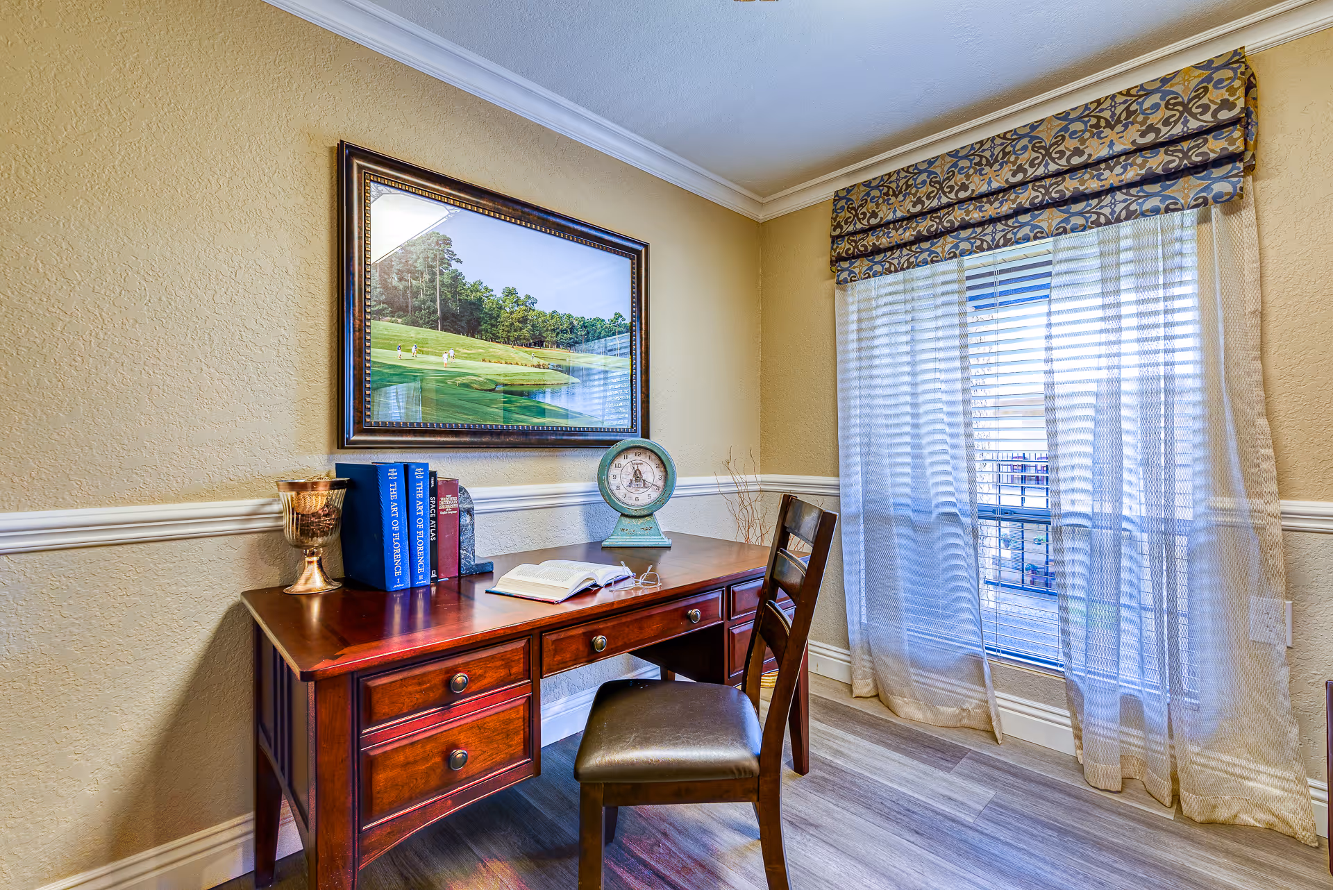A cozy study area in a senior living facility featuring a wooden desk with drawers, a matching wooden chair with a cushioned seat, an open book, a pair of reading glasses, a vintage scale, and two blue books on the desk. The room has beige textured walls with white trim, a large window with sheer curtains and a patterned valance, and a framed picture of a golf course hanging on the wall.