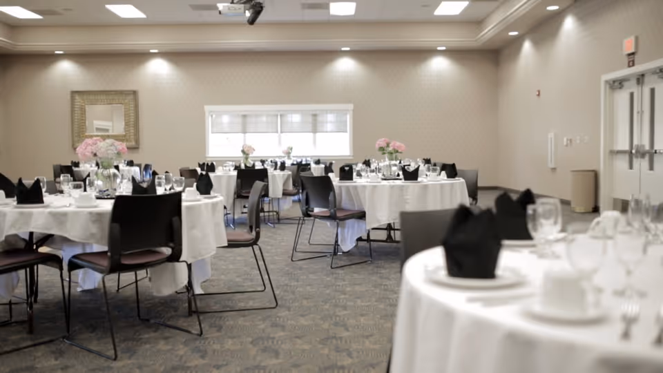 A dining room set up with round tables covered in white tablecloths, each table decorated with black folded napkins, glassware, white plates, and floral centerpieces with pink flowers. The room has beige walls, a carpeted floor, a window with blinds, and a framed mirror on one wall.