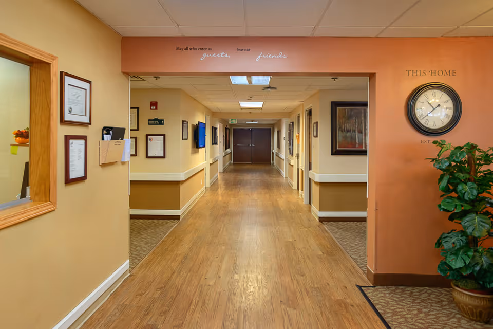A clean, well-lit hallway in a senior living facility with wood flooring and beige walls. The hallway is lined with framed certificates and artwork. There is a clock on the right wall above a potted plant, and a sign above the hallway entrance reads 'May all who enter as guests leave as friends.'