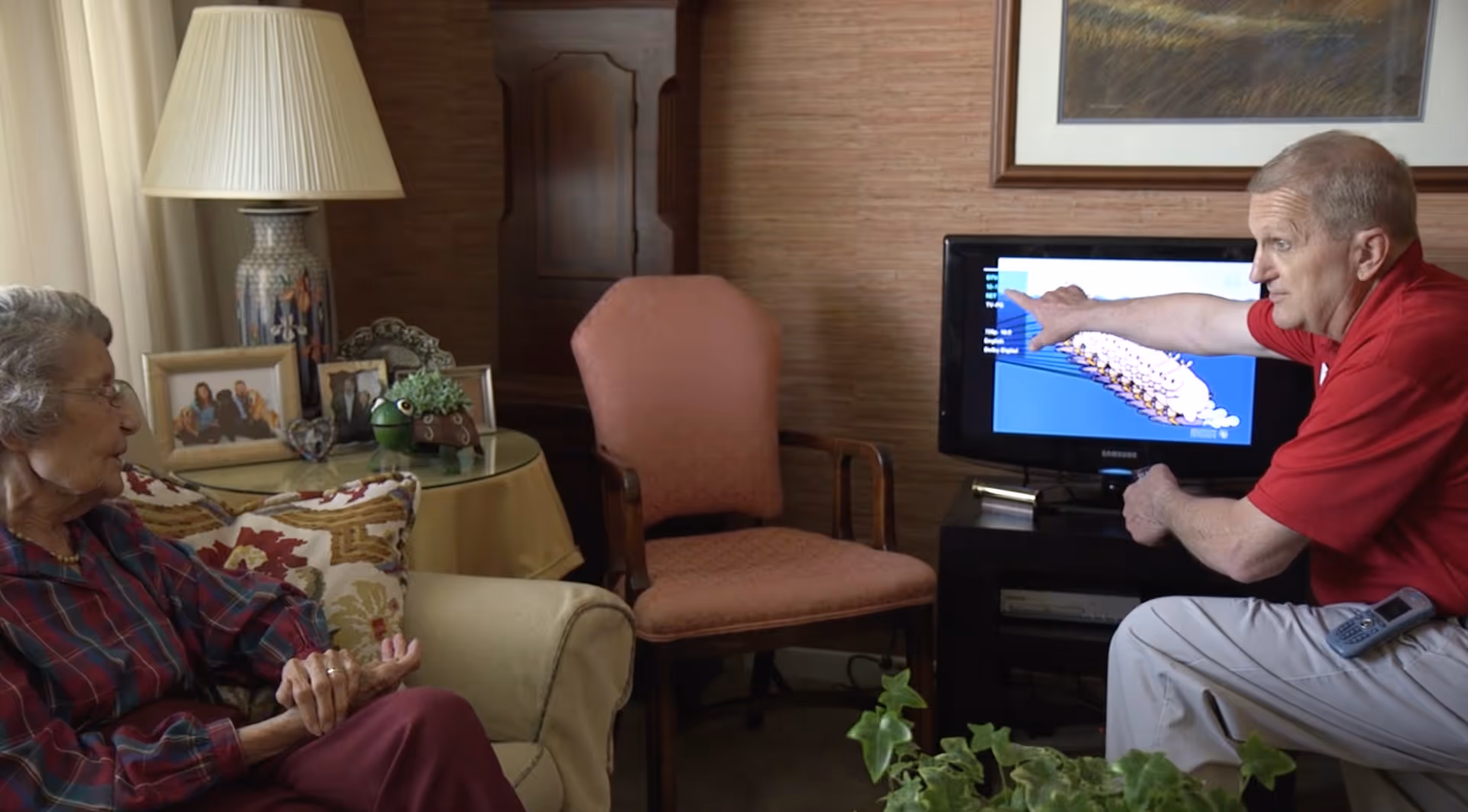 An elderly woman sitting on a couch with patterned pillows listens attentively to a man in a red shirt who is pointing at a diagram displayed on a television screen in a cozy living room setting with a lamp, framed photos, and a plant.