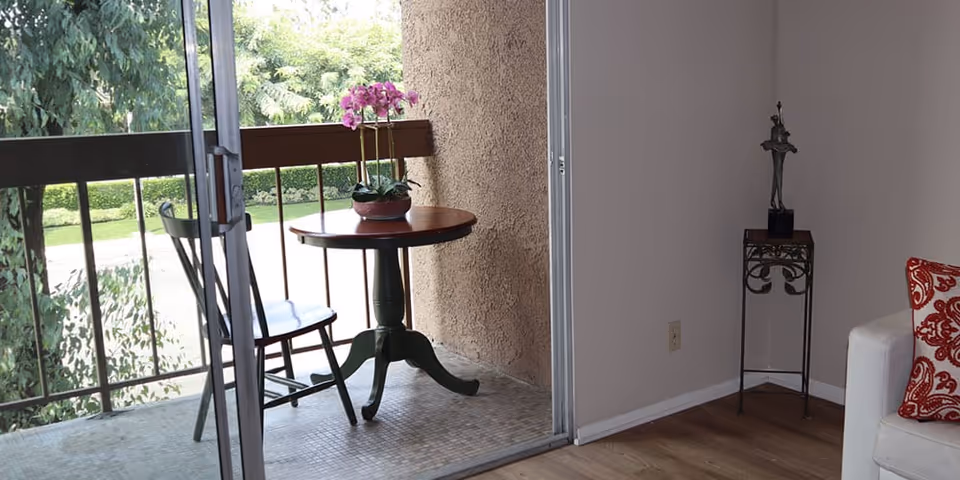A small balcony with a round wooden table and a single chair. On the table is a potted orchid with pink flowers. The balcony has a railing and overlooks green trees and bushes. Inside, part of a white couch with a red and white patterned pillow is visible, along with a small decorative stand holding a sculpture in the corner of the room.