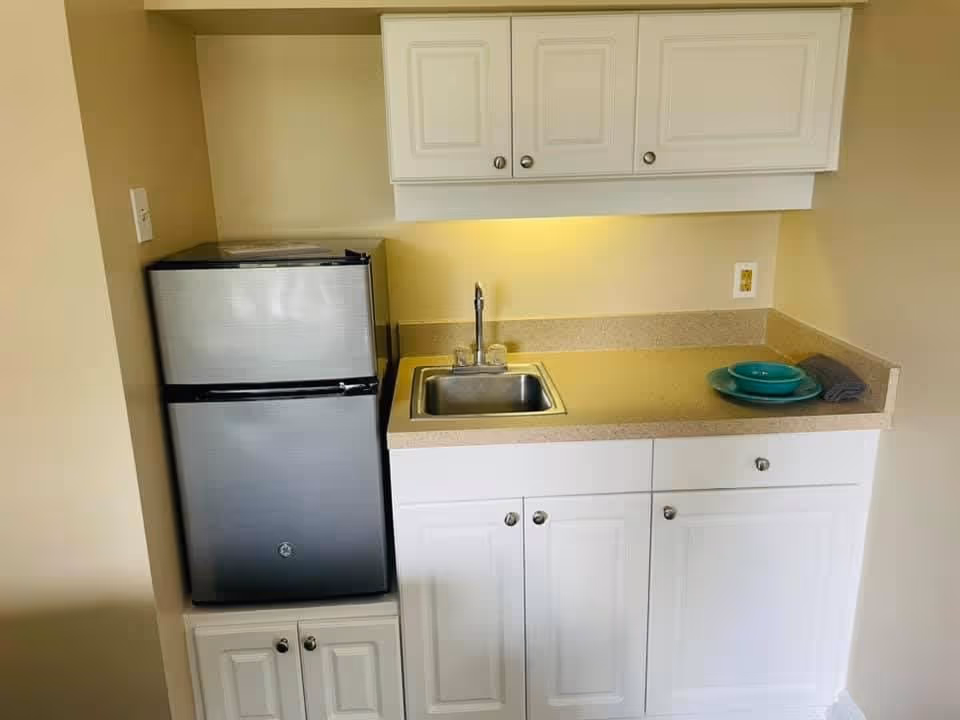 A small kitchenette area with a compact stainless steel refrigerator on the left, a countertop with a built-in sink and faucet in the center, white cabinets above and below the counter, and a set of turquoise plates and a folded dark cloth on the right side of the counter.