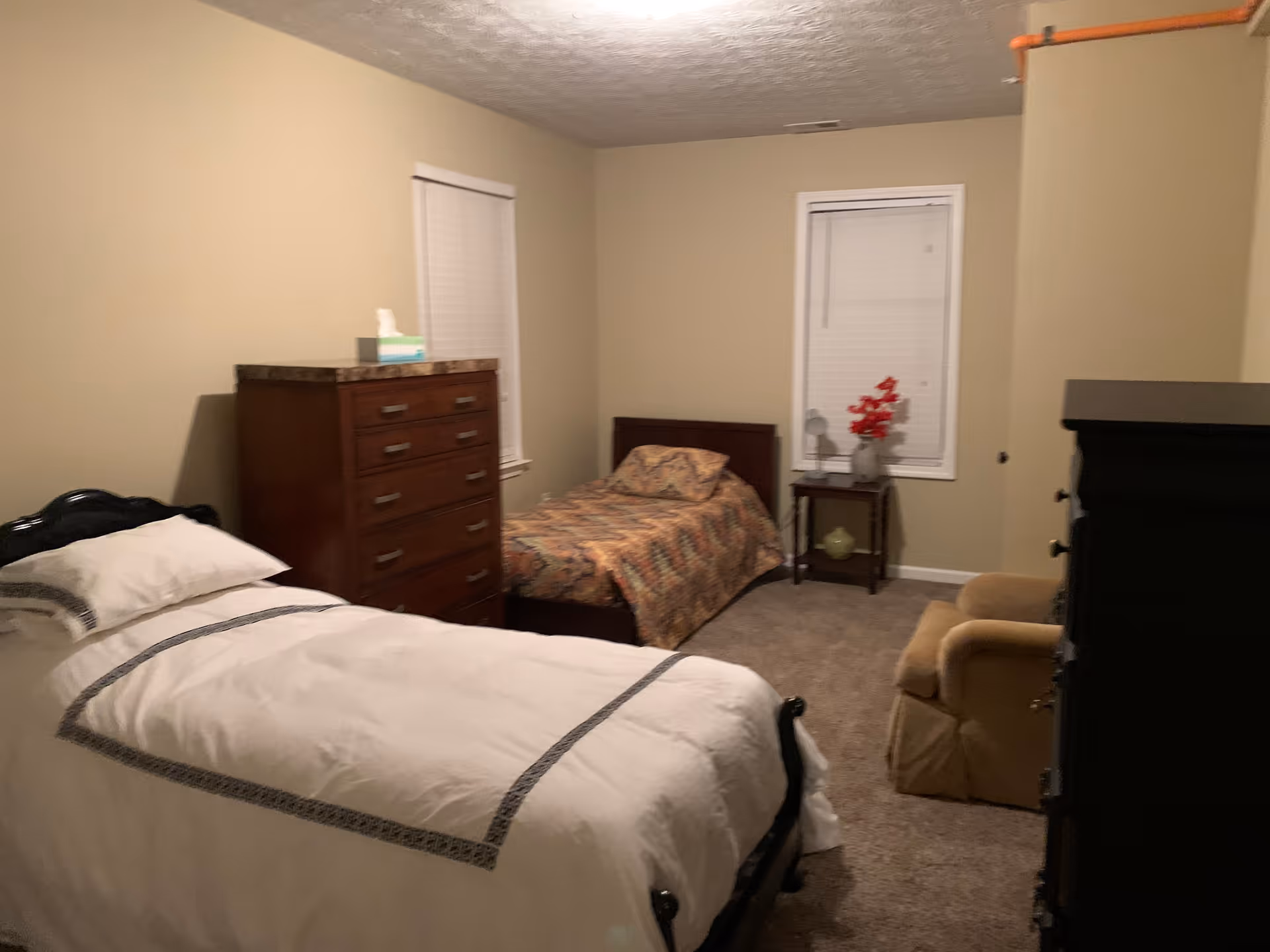 A bedroom with two single beds, one with white bedding and the other with patterned bedding. There is a wooden dresser between two windows with closed blinds, a small side table with a red flower arrangement, and a beige armchair in the corner.