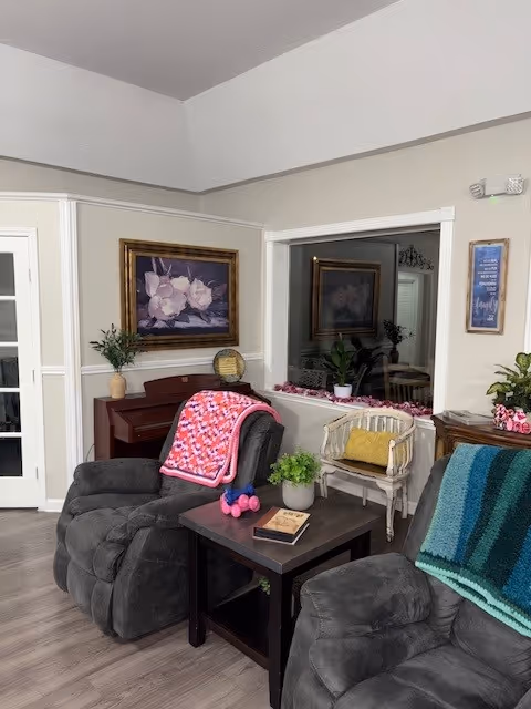 A cozy living room area with two gray recliner chairs, each draped with colorful knitted blankets. Between the chairs is a dark wooden side table holding a book, a small potted plant, and pink dumbbells. Behind the chairs is a small wooden piano with a vase of flowers and a decorative plate on top. A framed floral painting hangs on the wall above the piano. To the right, there is a white wooden chair with a yellow cushion and a large mirror reflecting part of the room. The walls are light-colored with white trim, and the floor is wood.