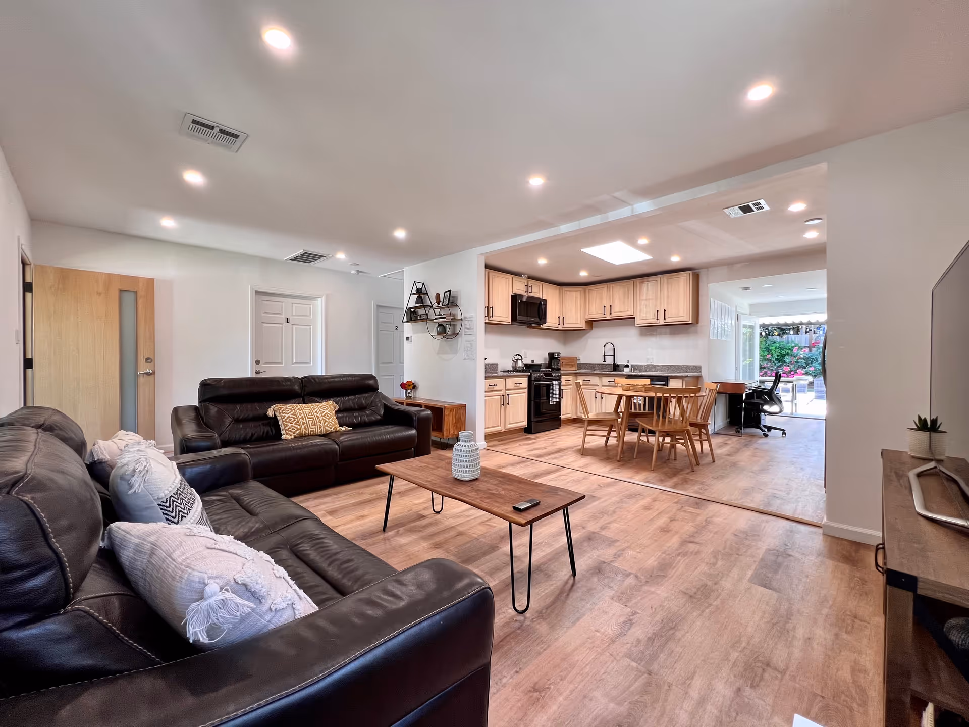 A bright and spacious senior living facility interior showing a living room with two black leather sofas adorned with white and patterned pillows, a wooden coffee table with a decorative vase, and a flat-screen TV on a wooden stand. The living room opens into a kitchen area with light wood cabinets, a black stove, microwave, and a wooden dining table with four chairs. Beyond the kitchen, there is a small office space with a desk and chair next to a glass door leading to an outdoor garden with blooming flowers.