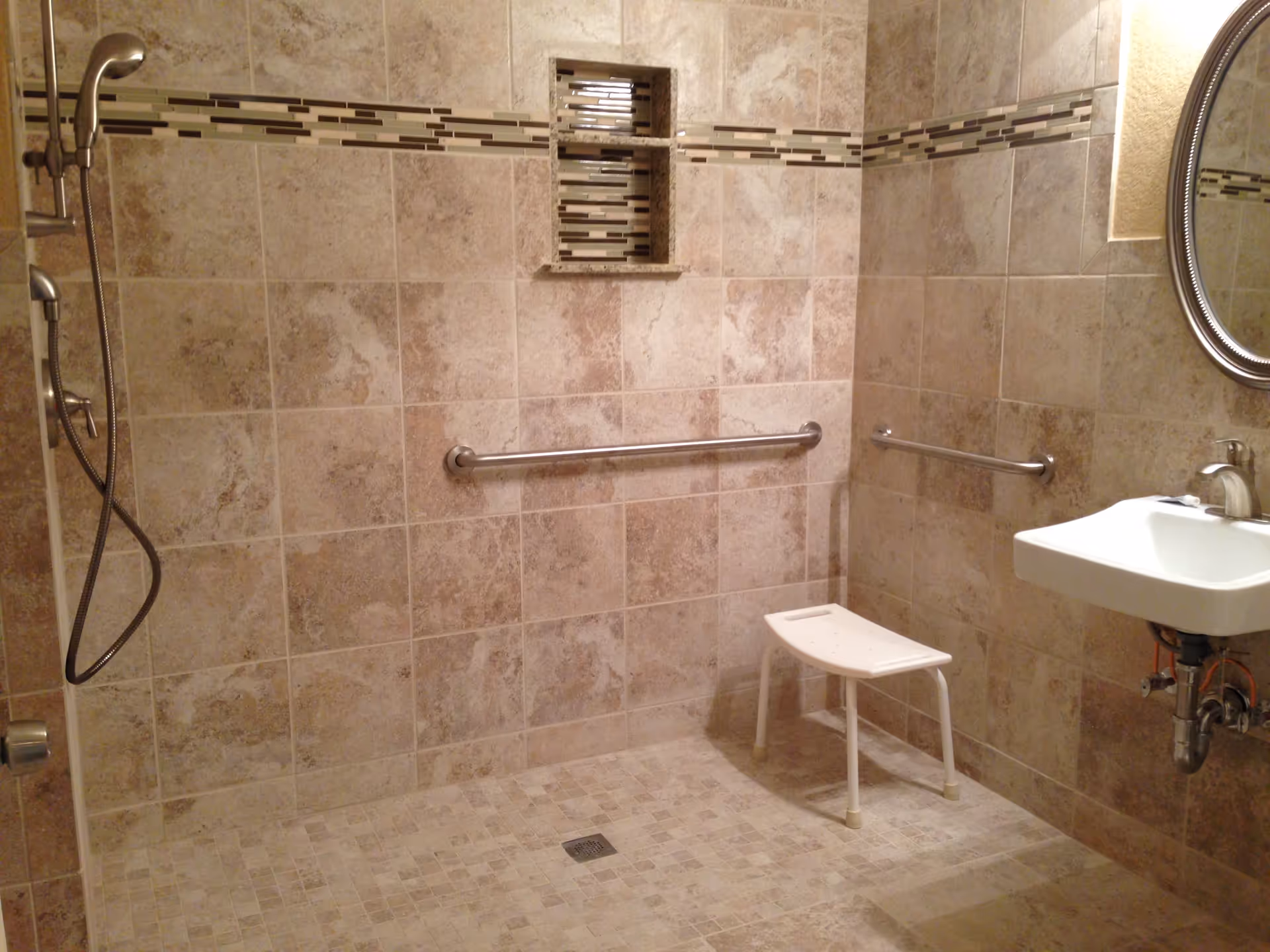 A tiled bathroom with a walk-in shower area featuring a handheld showerhead, two horizontal grab bars on the walls, a small white shower stool, a wall-mounted sink with a faucet, and a round mirror above the sink. The walls and floor are covered with beige and brown tiles, with a decorative horizontal tile strip running along the shower wall.
