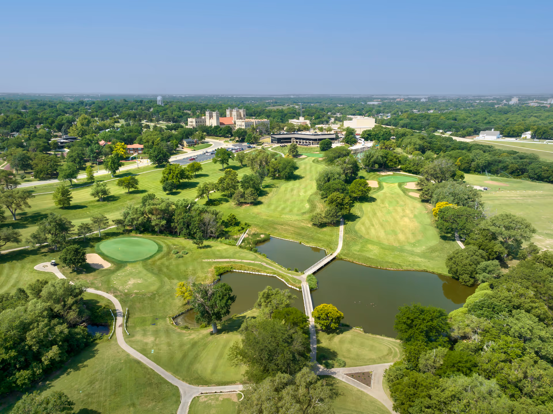 Aerial view of a green golf course with several fairways, sand bunkers, and water hazards. There are walking paths and bridges crossing the water. In the background, there are buildings and a parking lot surrounded by trees under a clear blue sky.