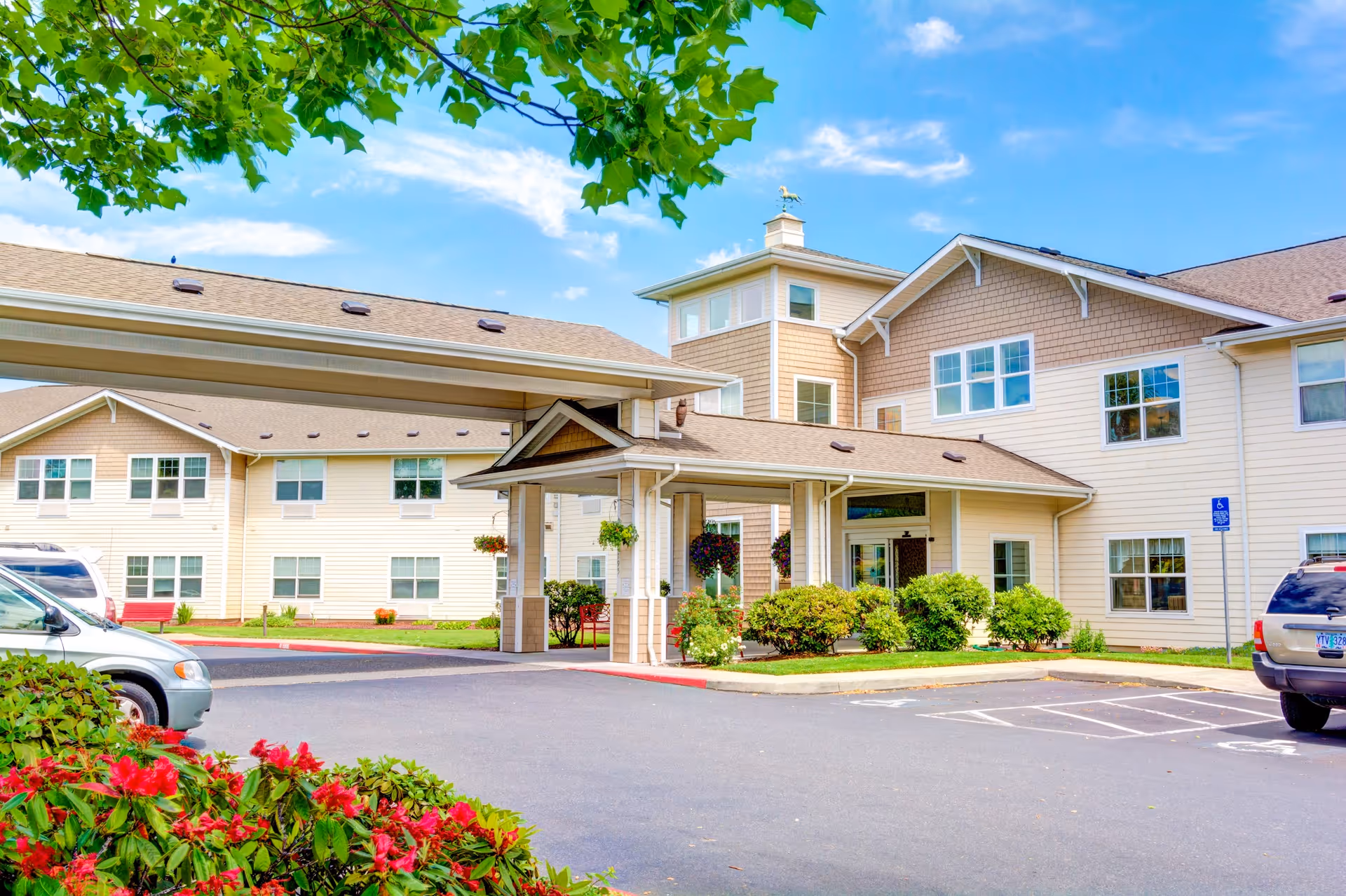 Exterior view of Hawks Ridge Assisted Living facility showing a beige multi-story building with a covered entrance, surrounded by greenery and parked cars under a blue sky with some clouds.