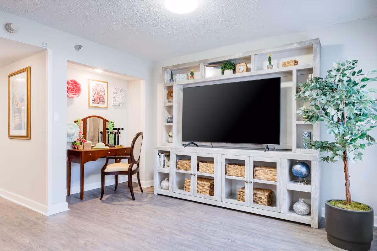 A bright living room area featuring a large flat-screen TV mounted on a white wooden entertainment center with shelves and wicker baskets. To the left, there is a small vanity table with a mirror, chair, and decorative items. A potted artificial tree is placed on the right side of the entertainment center. The room has light-colored walls and wood flooring.