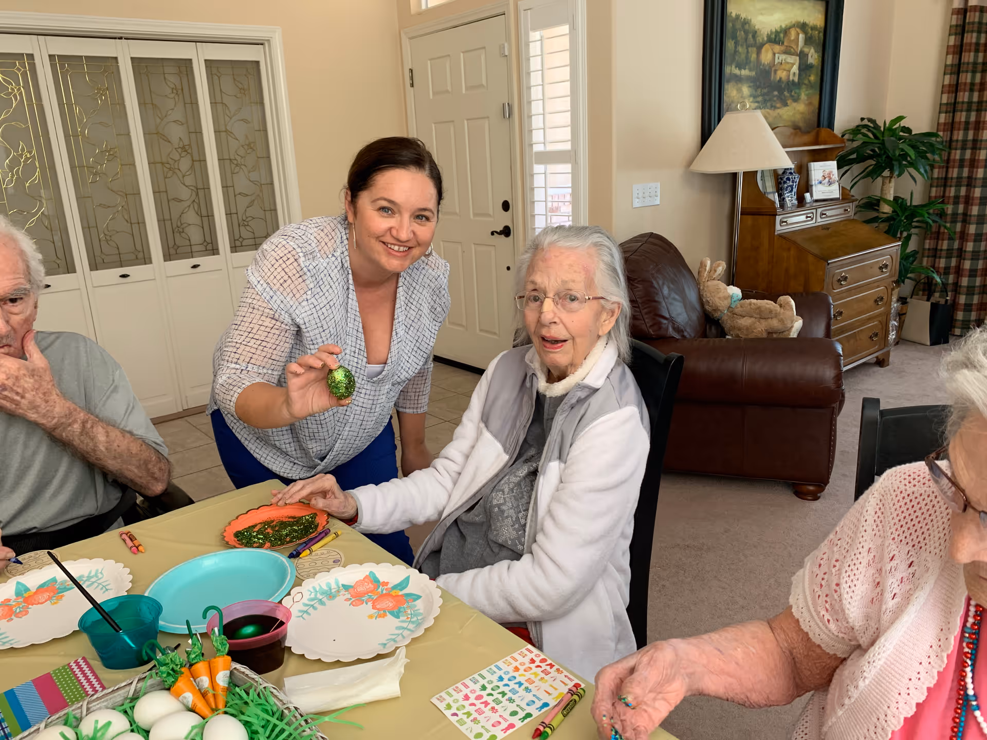 A group of elderly people and a caregiver are seated around a table in a living room area, engaging in an arts and crafts activity involving decorating Easter eggs. The caregiver is smiling and holding a decorated green egg. The room has a brown leather couch, a wooden cabinet, a lamp, and a painting on the wall.