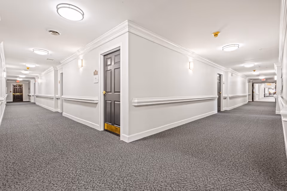 A clean, well-lit hallway in a senior living facility with white walls, gray carpet, and multiple dark brown doors. The hallway features ceiling lights, wall sconces, and handrails along the walls. The corridor extends in two directions from a corner, with exit signs visible at both ends.