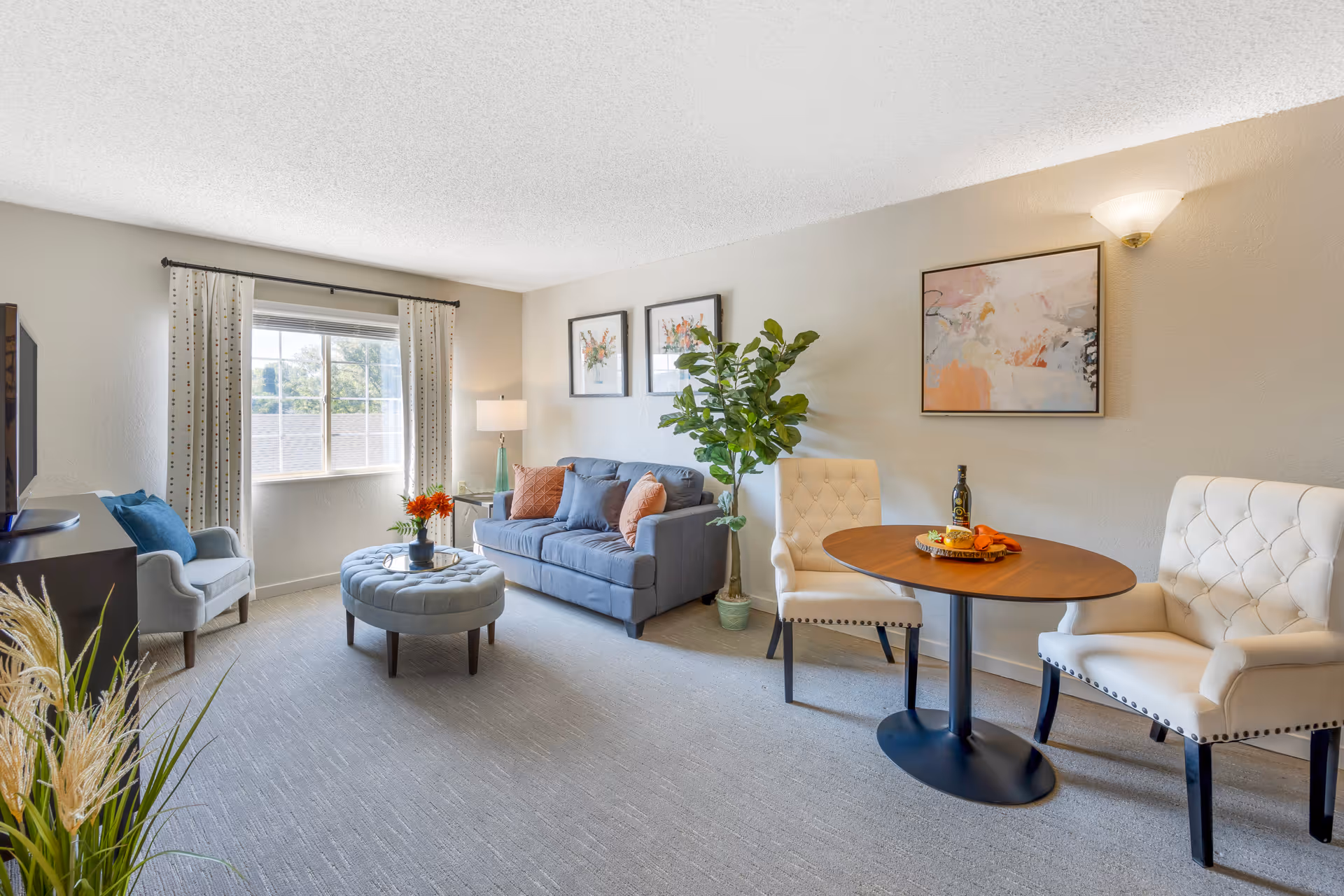 A bright and cozy living room with a large window letting in natural light. The room features a blue sofa with orange and blue cushions, a round tufted ottoman, a small side table with a lamp, two cream-colored upholstered chairs around a small wooden table with a bottle and fruit tray, a potted plant, and framed artwork on the walls.