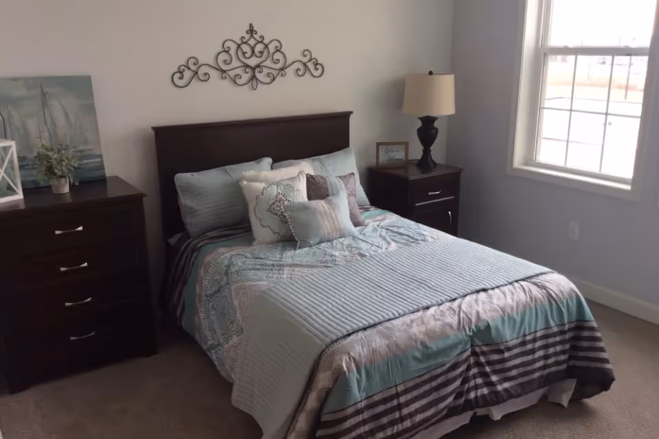 A neatly made bed with a patterned blue and gray comforter and multiple pillows in a bedroom. The room has a dark wooden headboard, matching nightstand with a lamp, and a dresser with decorative items including a small plant and a painting of sailboats. A window on the right side lets in natural light.