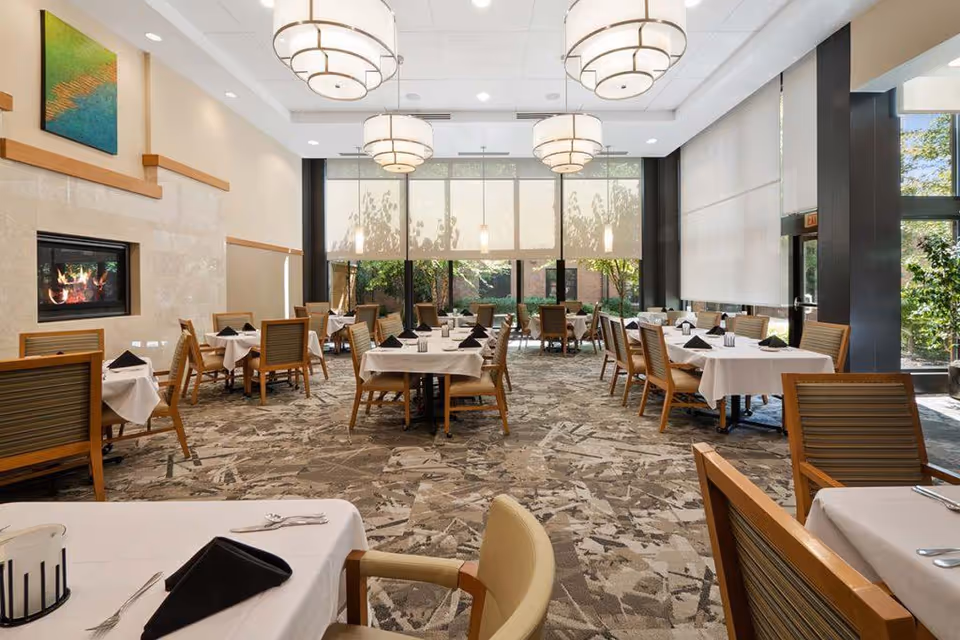 A spacious dining room with multiple tables covered in white tablecloths, each set with black folded napkins and silverware. The room features large windows with roller shades, letting in natural light and providing a view of greenery outside. There are modern pendant lights hanging from the ceiling and a fireplace on the left wall beneath a colorful abstract painting. The floor is covered with a patterned carpet, and wooden chairs with cushions surround the tables.