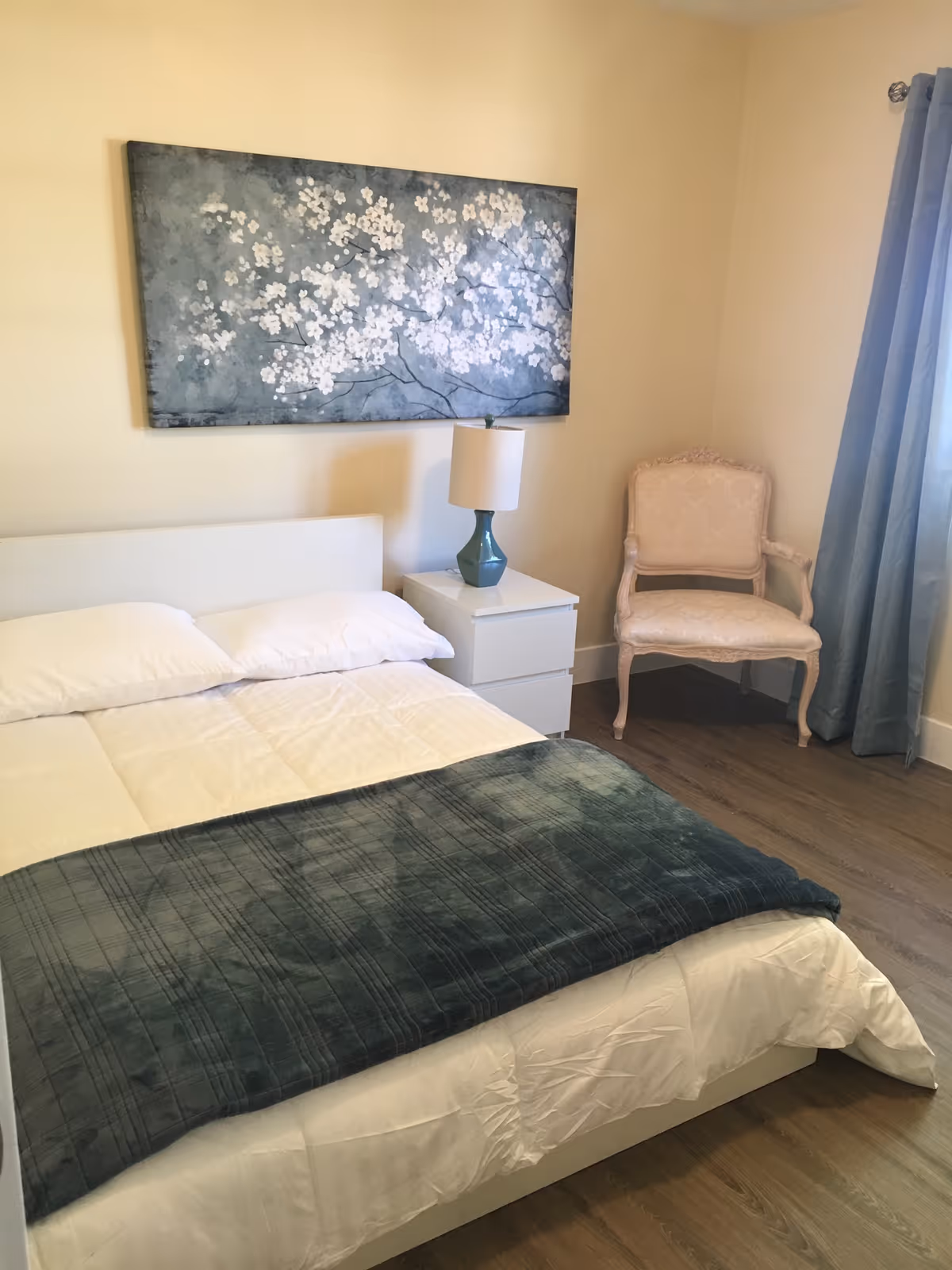 A neatly made bed with white bedding and a dark green blanket at the foot. Next to the bed is a white nightstand with a blue lamp. On the wall above the bed is a large painting of white flowers on a blue-gray background. To the right of the nightstand is an upholstered armchair with a light pink floral pattern. A window with blue curtains is partially visible on the right side of the room. The floor is wooden.