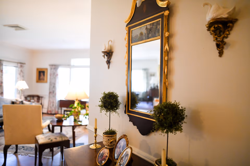 Interior view of a living room area with a decorative mirror on the wall, two small potted topiary plants, framed photos, and a candle holder on a wooden table. In the background, there are chairs, a coffee table, lamps, and windows with floral curtains allowing natural light into the room.