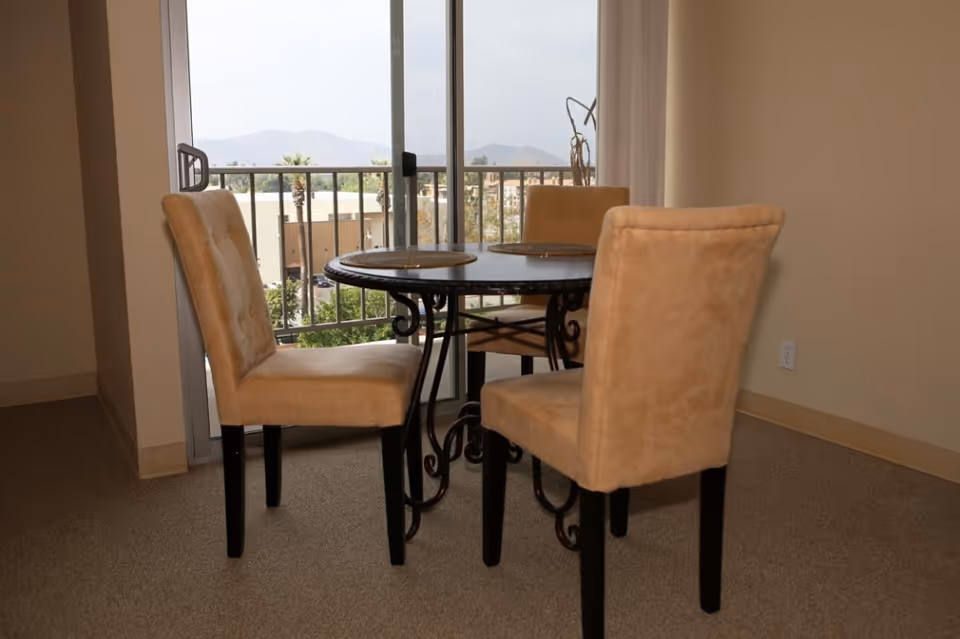A small round dining table with a black metal base and a dark tabletop, surrounded by three beige upholstered chairs. The table has three round placemats on it. Behind the table is a sliding glass door leading to a balcony with a view of buildings, palm trees, and distant mountains under a cloudy sky.