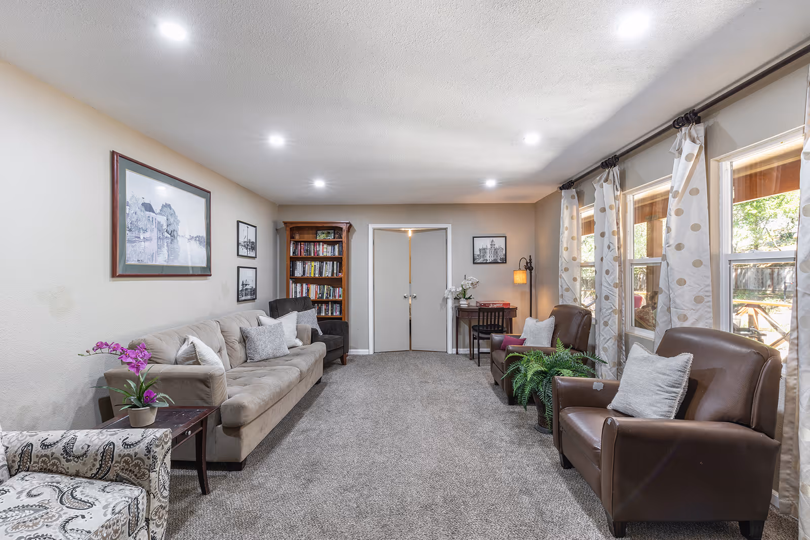 Carpeted communal living room with a sofa, armchairs, bookshelf, side tables, plants, and windows with curtains.