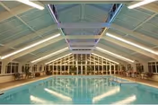 Indoor swimming pool with clear blue water under a high vaulted ceiling with large windows at the far end. Poolside chairs and tables line both sides of the pool area.