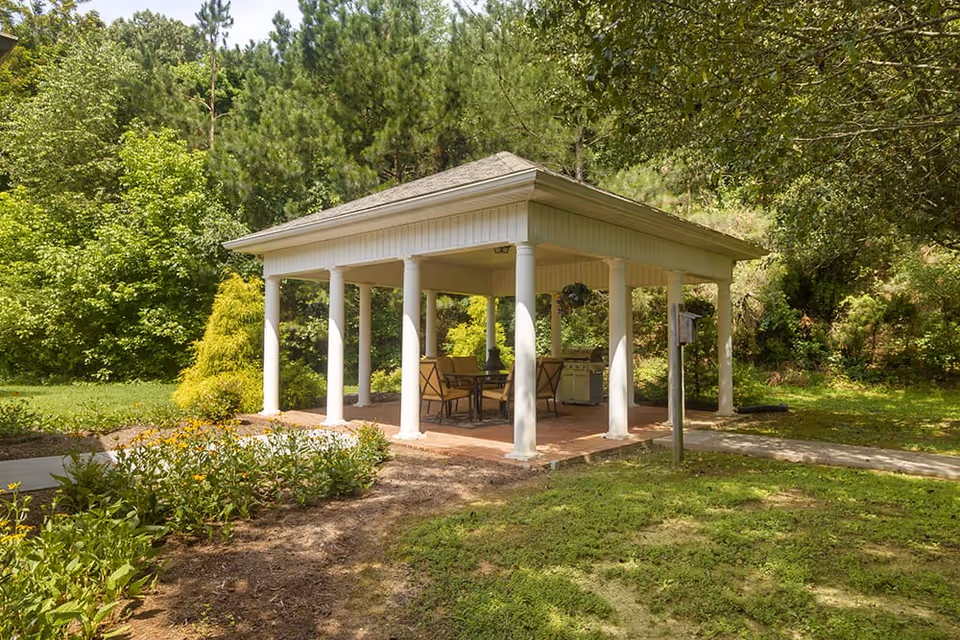 A white gazebo with a shingled roof and multiple columns stands in a lush green garden area. Inside the gazebo, there is a table with chairs and a grill. Surrounding the gazebo are trees, bushes, and a pathway leading to it.