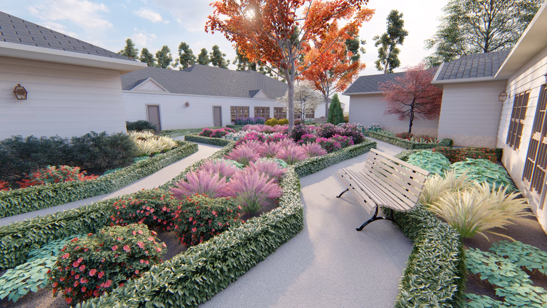 A landscaped outdoor garden area with a paved walkway, surrounded by various bushes, flowering plants, and trees with autumn-colored leaves. There is a wooden bench along the path, and white buildings with gray roofs are visible in the background under a partly cloudy sky.