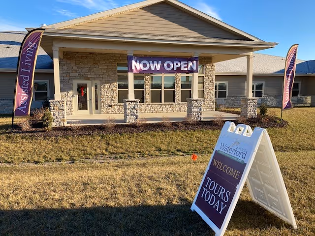 Front exterior of the Waterford Place assisted living building with a 'NOW OPEN' banner, flags, and an A-frame sign advertising tours.
