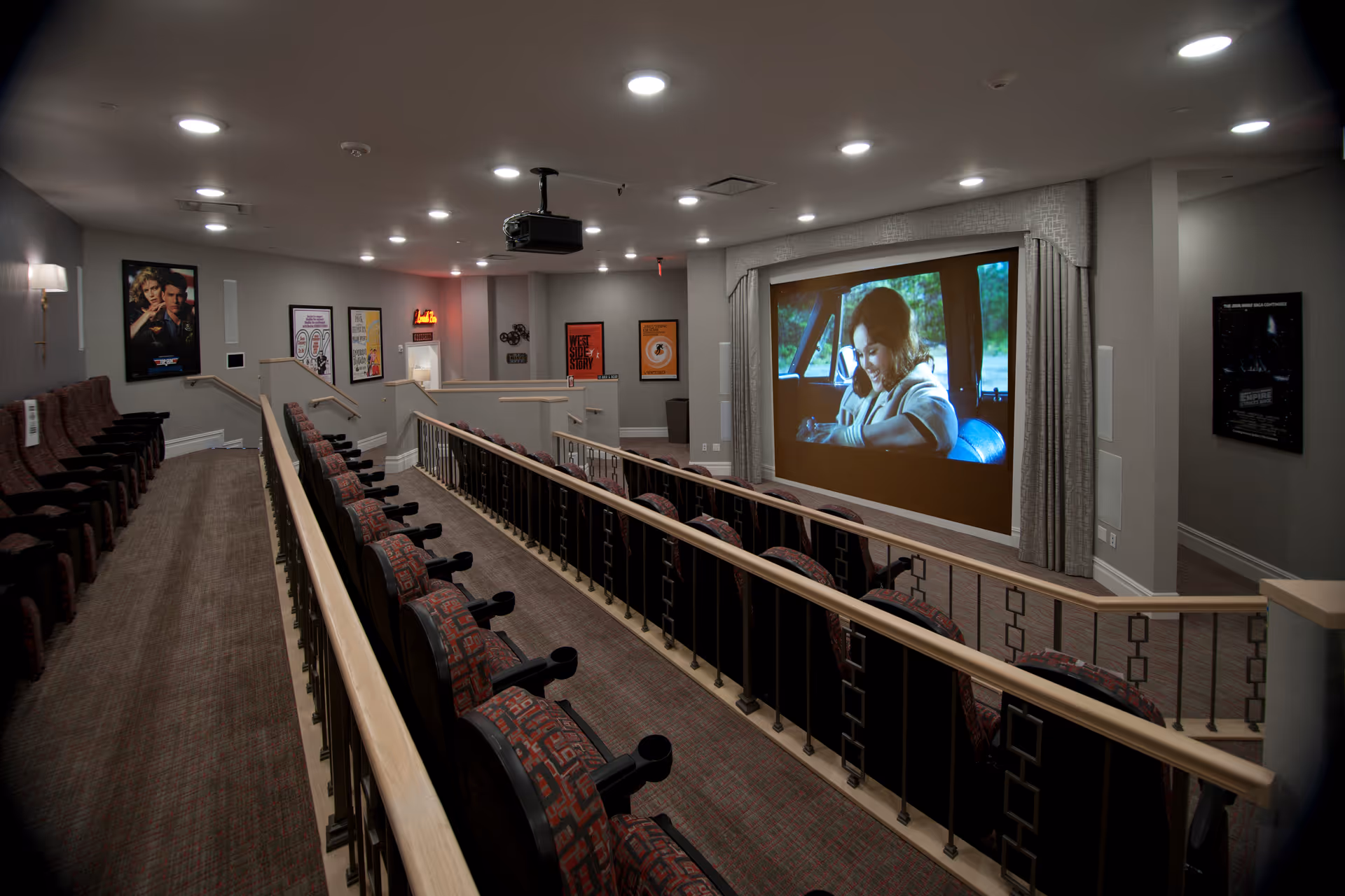 Interior view of a small theater room with tiered seating and a large screen showing a woman sitting in a car. The room has carpeted floors, framed movie posters on the walls, and ceiling lights. There is a projector mounted on the ceiling.